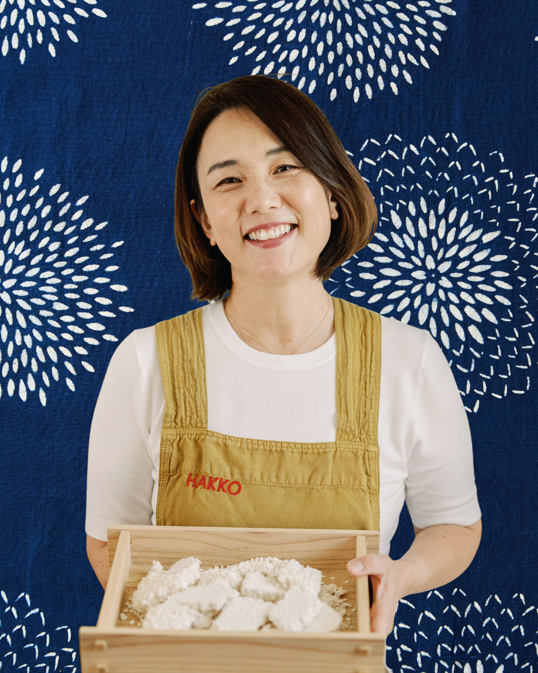 A smiling woman in a white shirt and yellow apron holding a wooden tray with rice flour, standing against a blue backdrop with white firework-like patterns.
