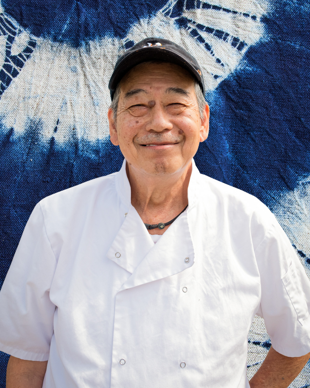 Smiling older man wearing a chef's coat and black hat, standing in front of a blue and white fabric backdrop.