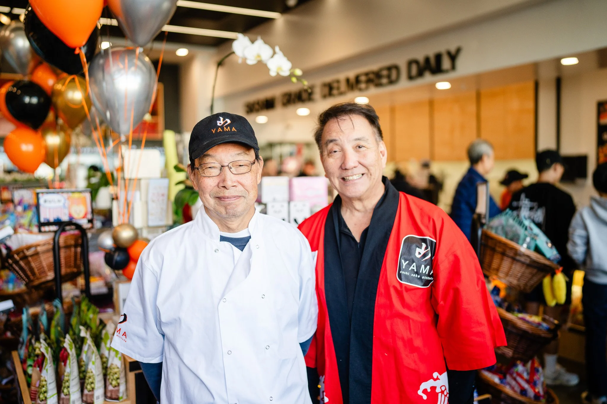 Two men stand smiling in a vibrant store filled with balloons and colorful products. One wears a chef's coat, the other a red jacket.