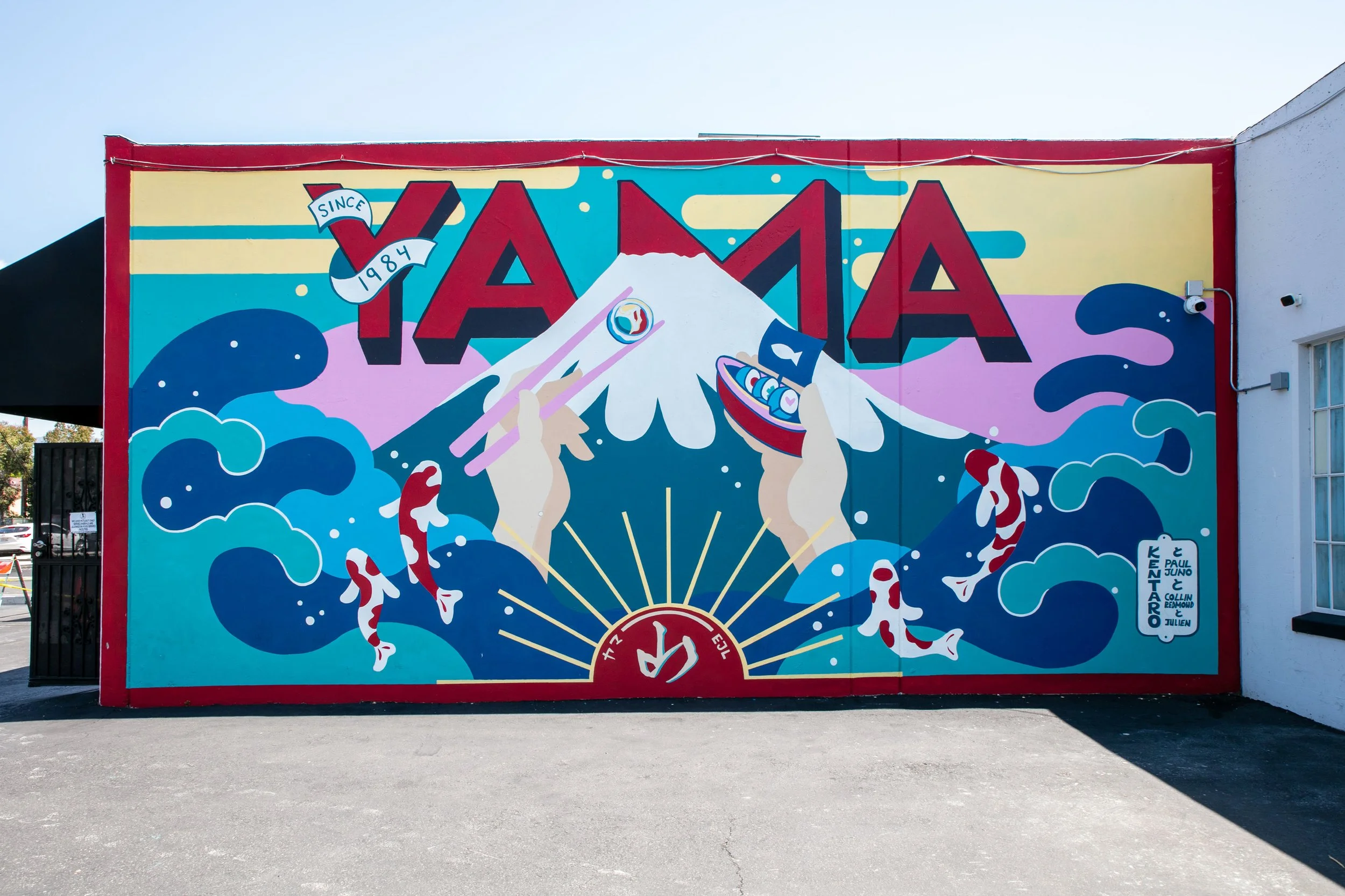 Colorful mural with the word YAMA written above a stylized mountain peak, surrounded by abstract waves, koi fish, and hands holding a fish and a torii gate, with the text 'Since 1984.'