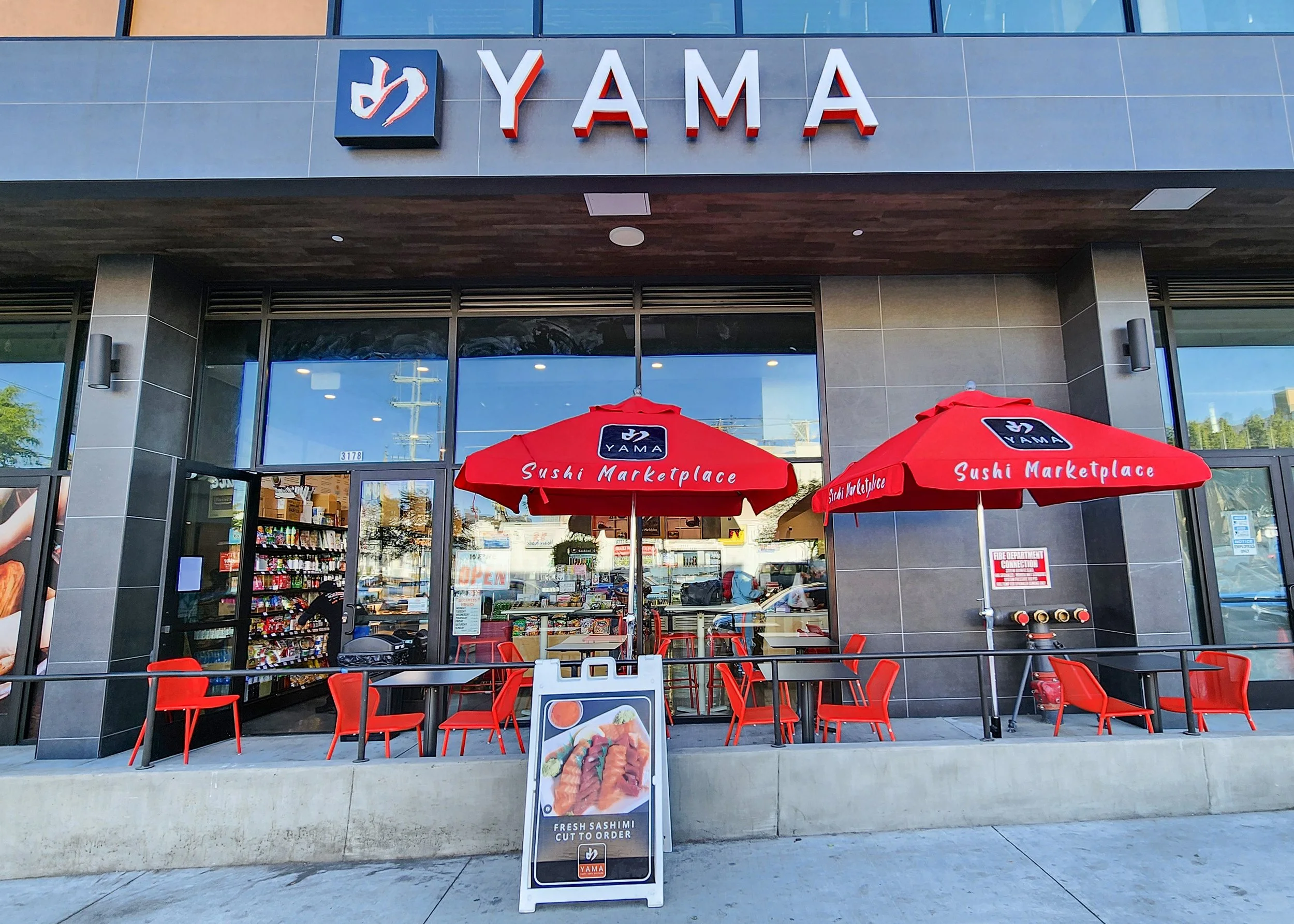 Exterior of YAMA sushi marketplace with red umbrellas and red chairs outside, a signboard displaying fresh sashimi, and large glass windows showing the interior of the restaurant.