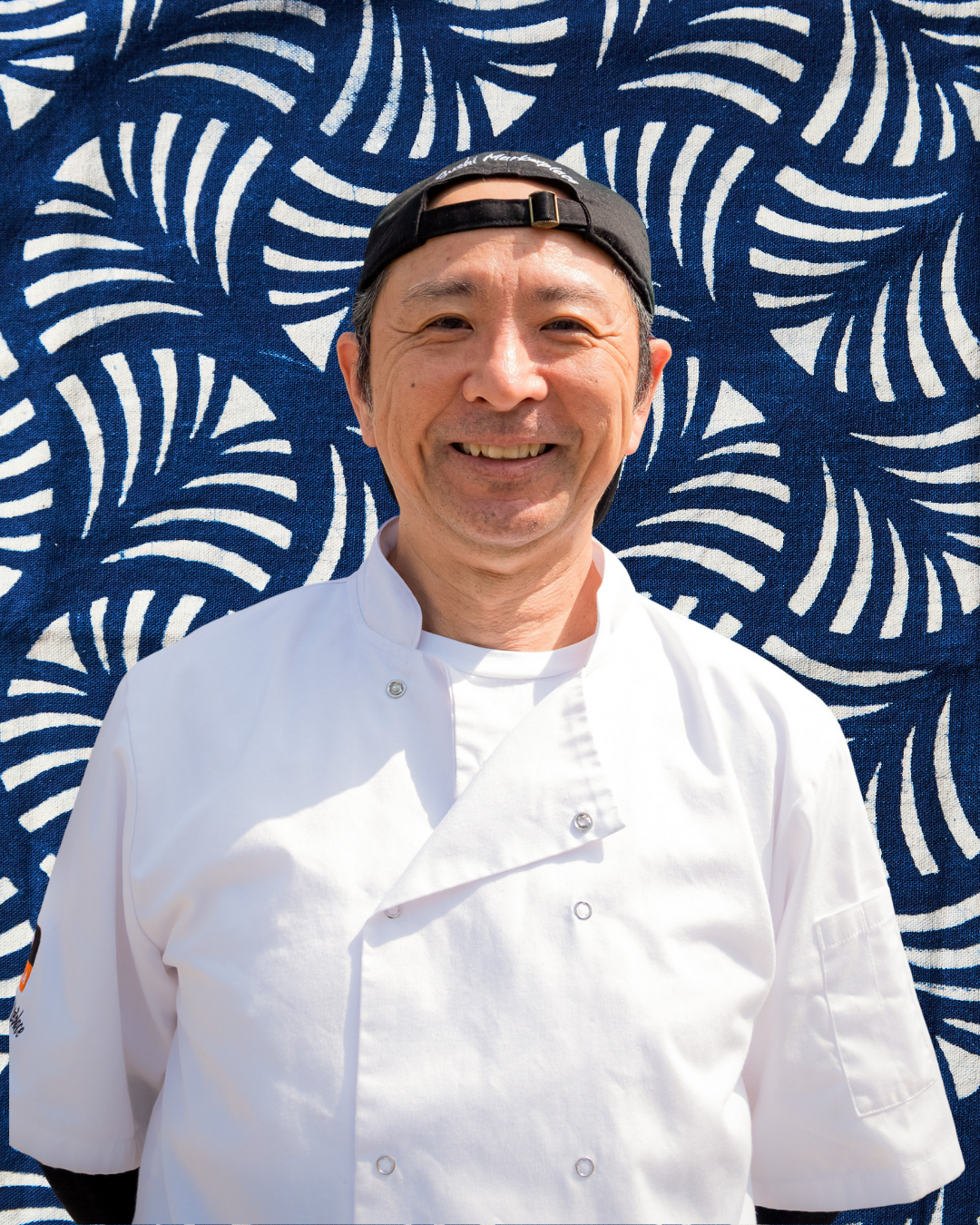 Smiling chef in white uniform and black cap standing in front of a blue and white patterned background.