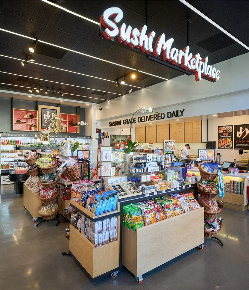 Interior of a sushi marketplace with shelves of snacks, a sign reading 'Sashimi Grade Delivered Daily,' and a staff member working behind the counter.