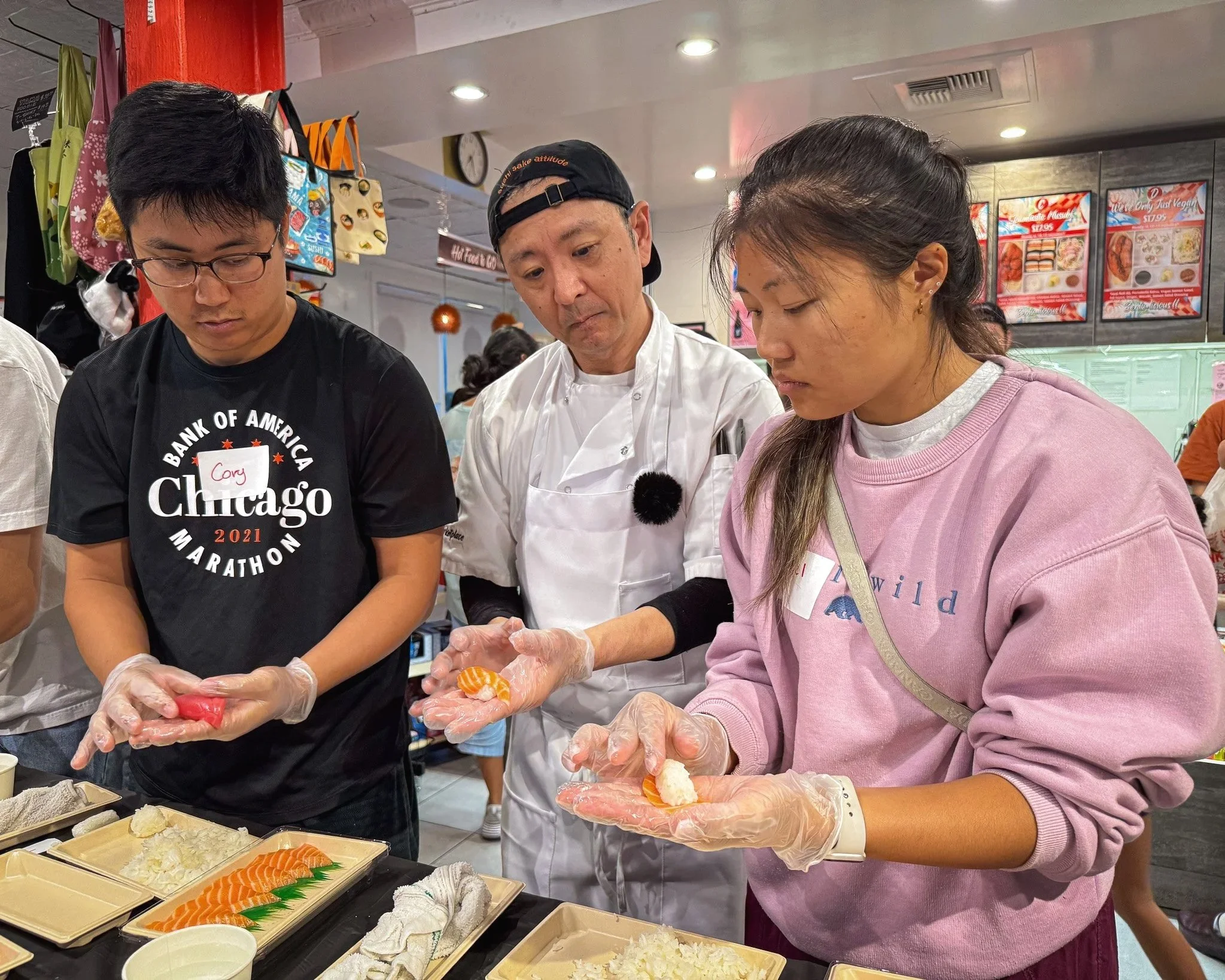 Three people are preparing sushi in a cooking class. A chef guides two participants, one wearing a black shirt and the other in a pink sweatshirt.