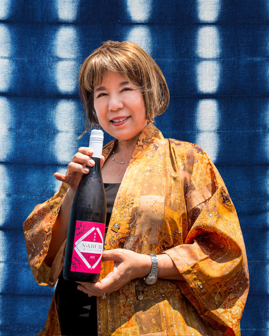 A woman with short brown hair holding a dark bottle with a pink label that says 'NARUMI CRAFT SAKE Jazz' against a blue fabric background with white circular patterns. She is smiling and wearing a colorful, patterned kimono-style jacket.