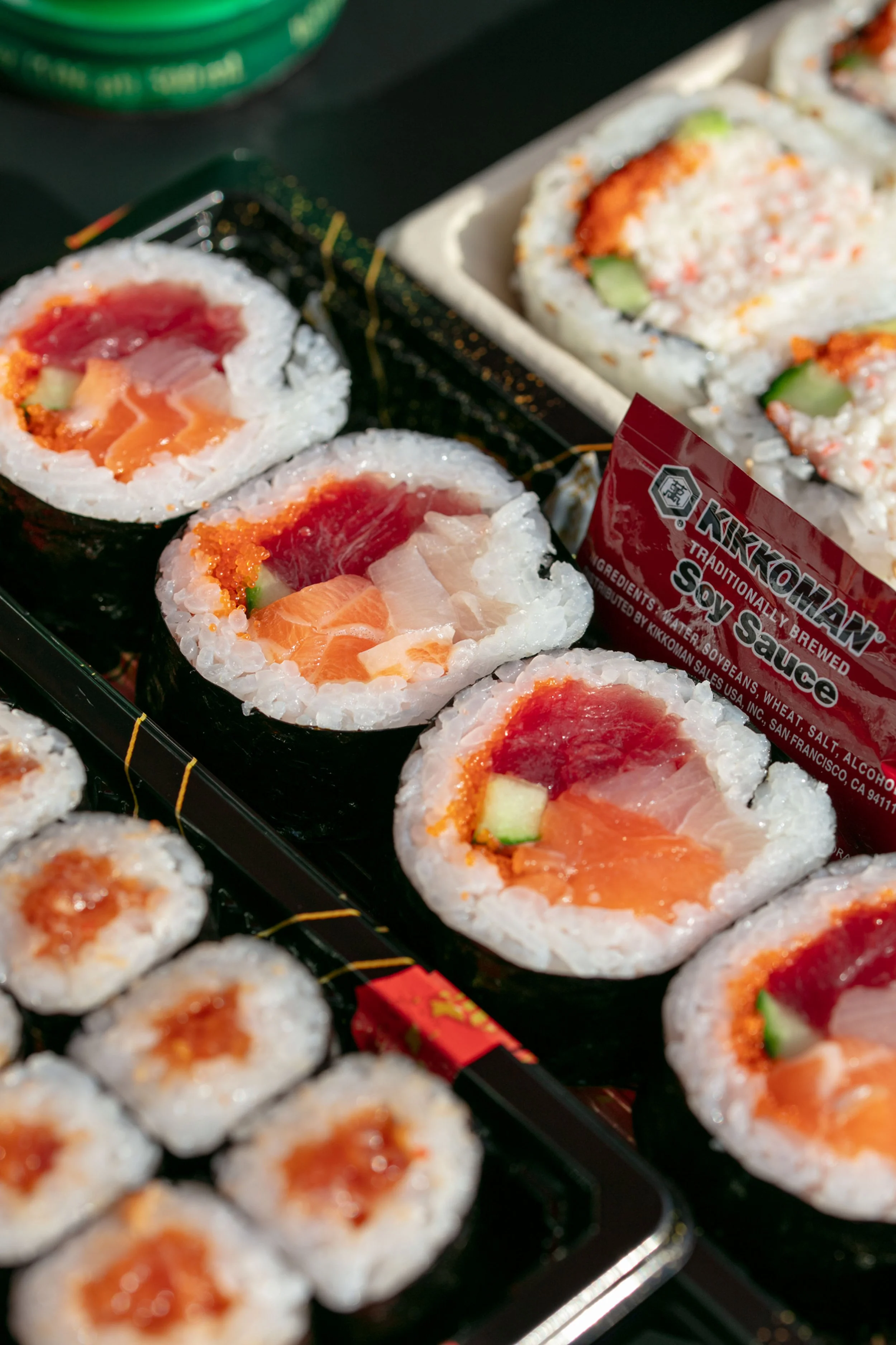 Assorted sushi rolls including salmon, tuna, and cucumber, displayed in black trays with a red soy sauce packet.